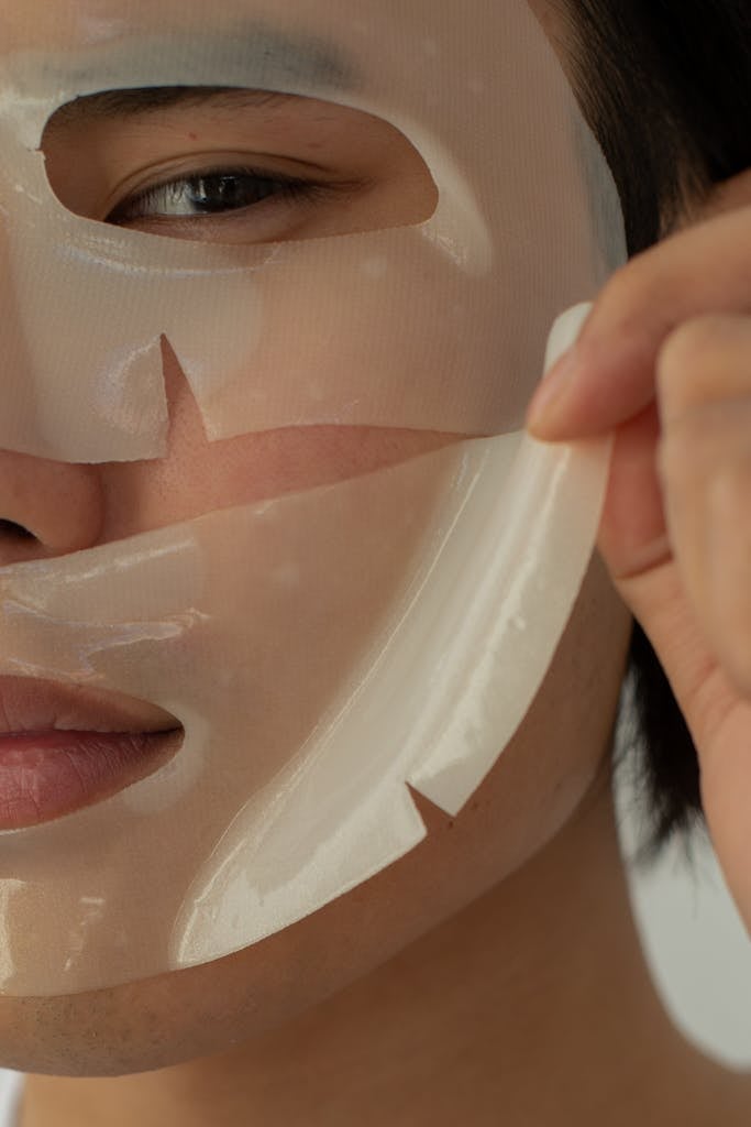 Crop unrecognizable Asian male model looking at camera while taking off moisturizing facial mask during skincare routine in light studio